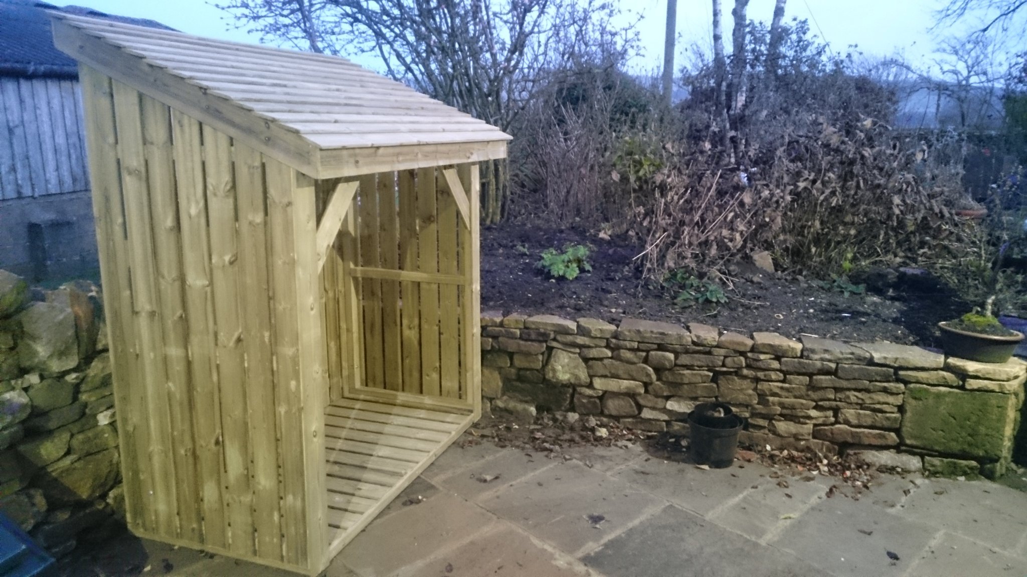 Open fronted timber log store against a dry stone wall